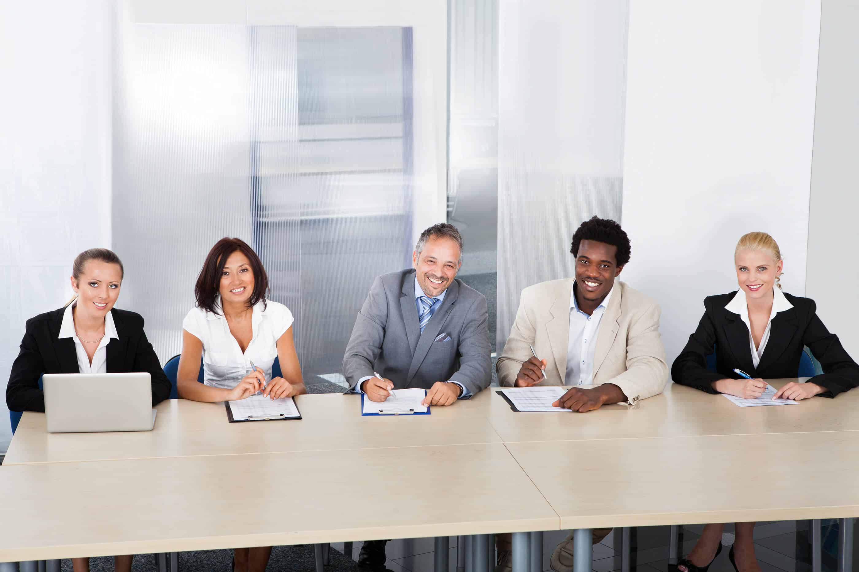A group of 5 people of all different ethnicities dressed in business suits. There are 2 males and 3 females. They are all sitting down at a long table, looking like they are evaluating something. 4 of them have a clipboard and a pen. The woman on the left end has a laptop open in front of her.