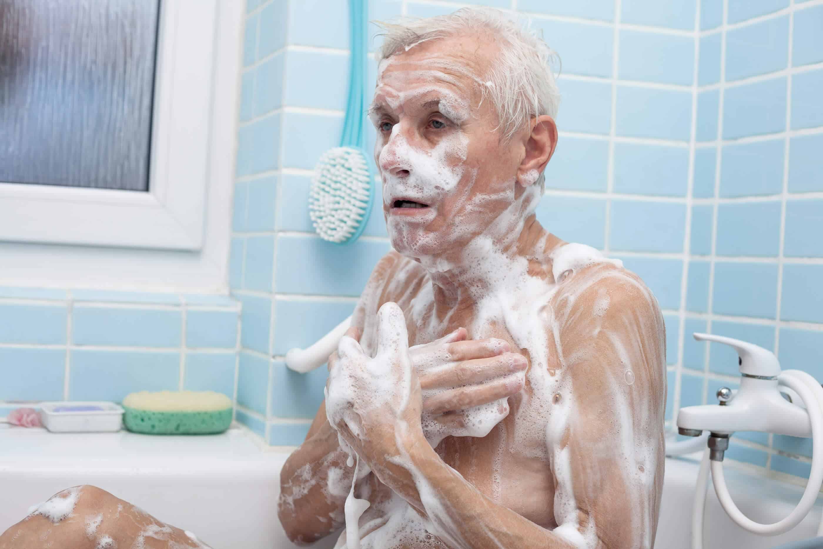 An Old Man with white hair is in a bathtub, and is completely covered in soap bubbles. He is looking off to the side. The background is blue tile with a sponge and long blue scrubbing brush next to the man.
