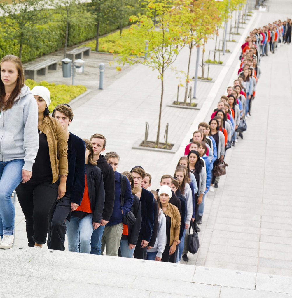A long line of people standing very close to one another. Some trees are seen in the background. The line is going up a flight of stairs.