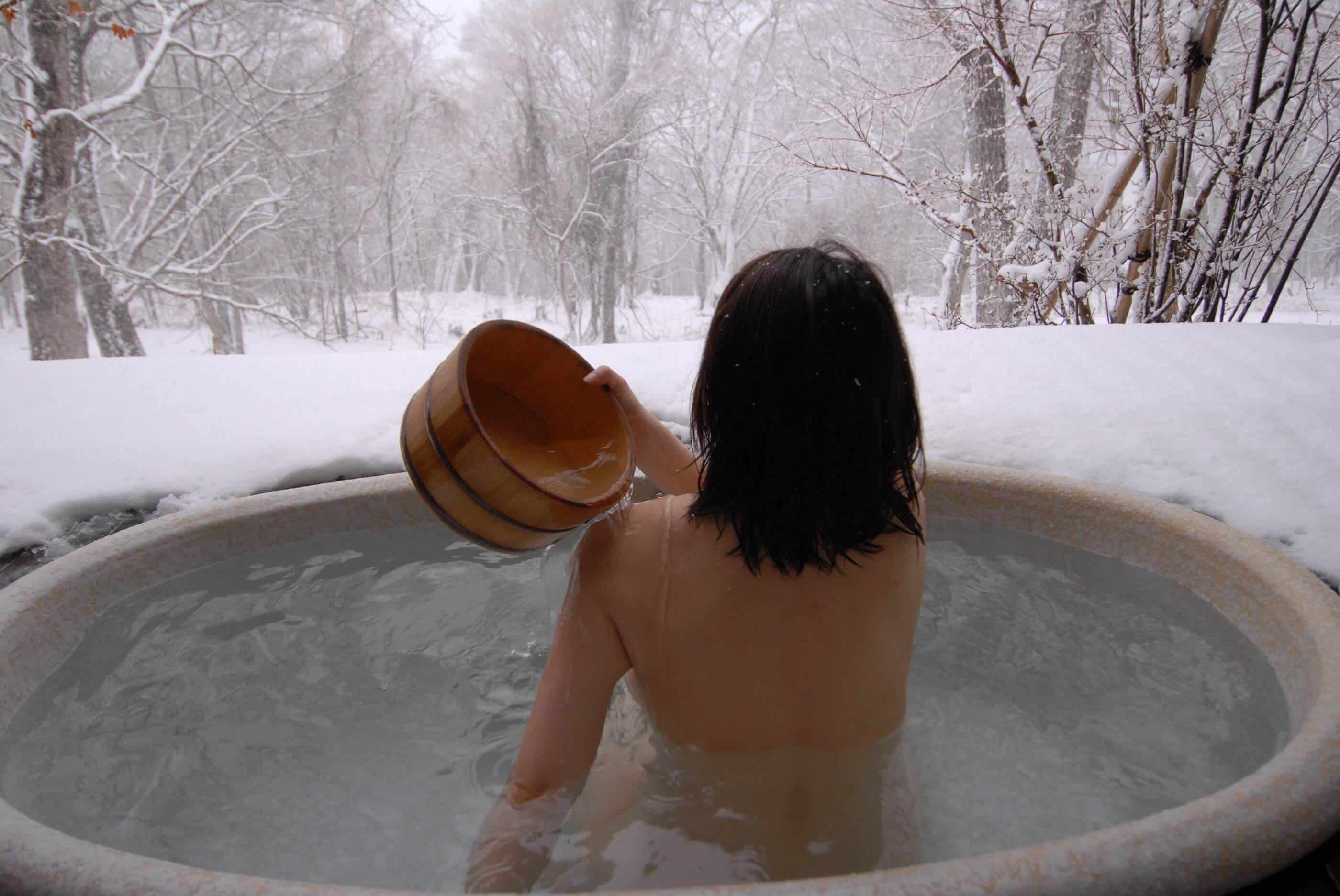 A woman with black hair is soaking in a Japanese Rotenburo (open air bath) while looking at the snow and trees all around her.