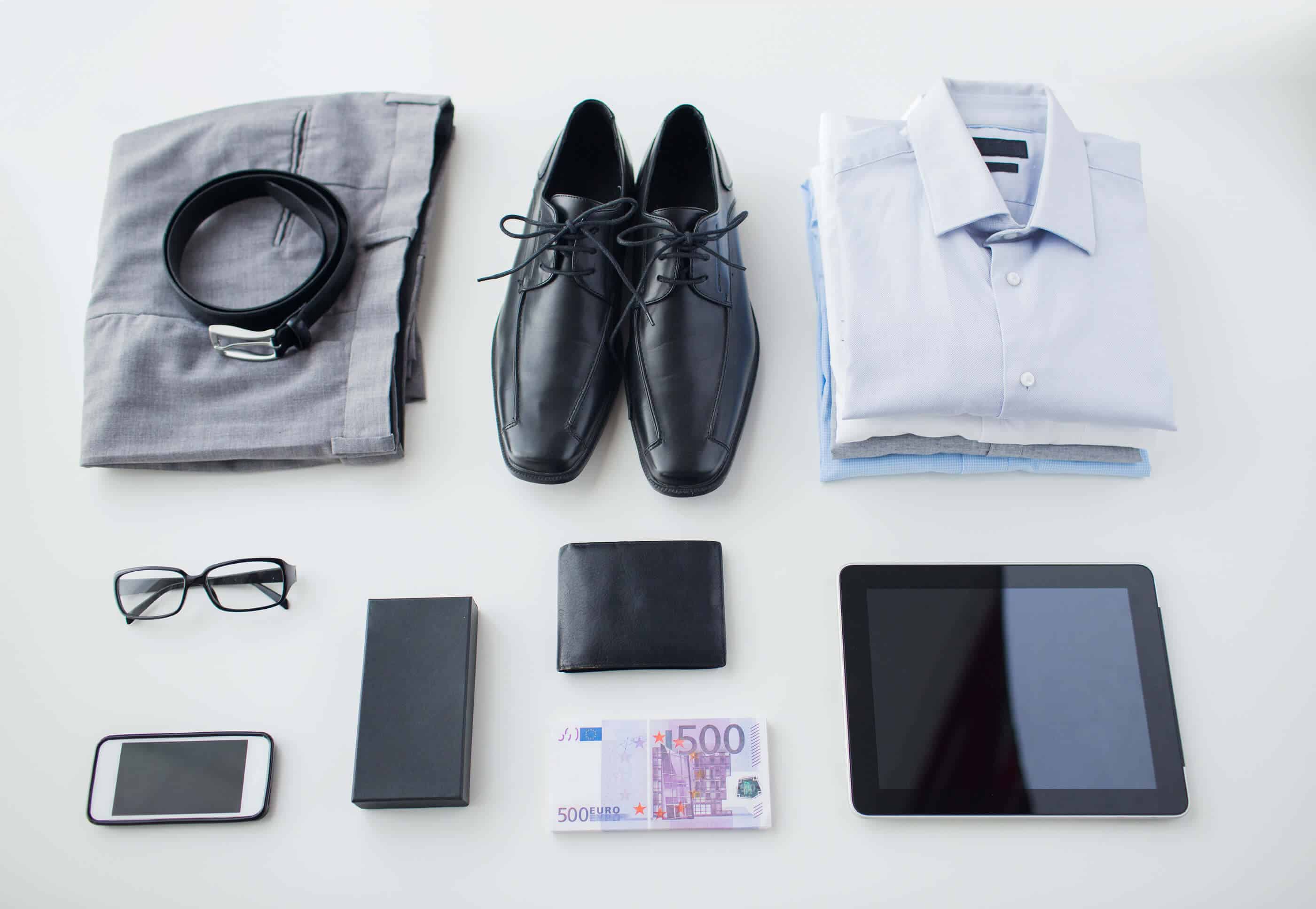 A folded pair of grey pants with a black belt on top, a pair of black dress shoes, three folded button up shirts, a pair of glasses, a smartphone, a wallet, a longer pocket wallet, a electronic tablet and cash on a white surface.