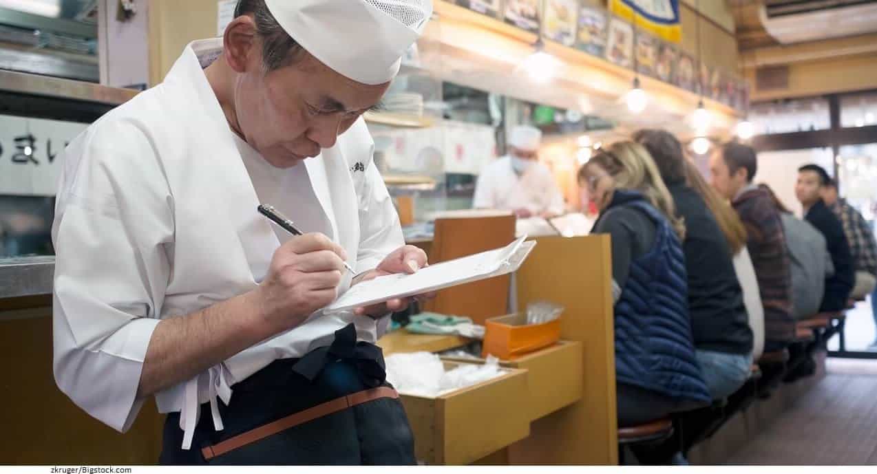 An older Asian man wearing a cooks uniform is writing something down on a restaurant order sheet inside of a restaurant.