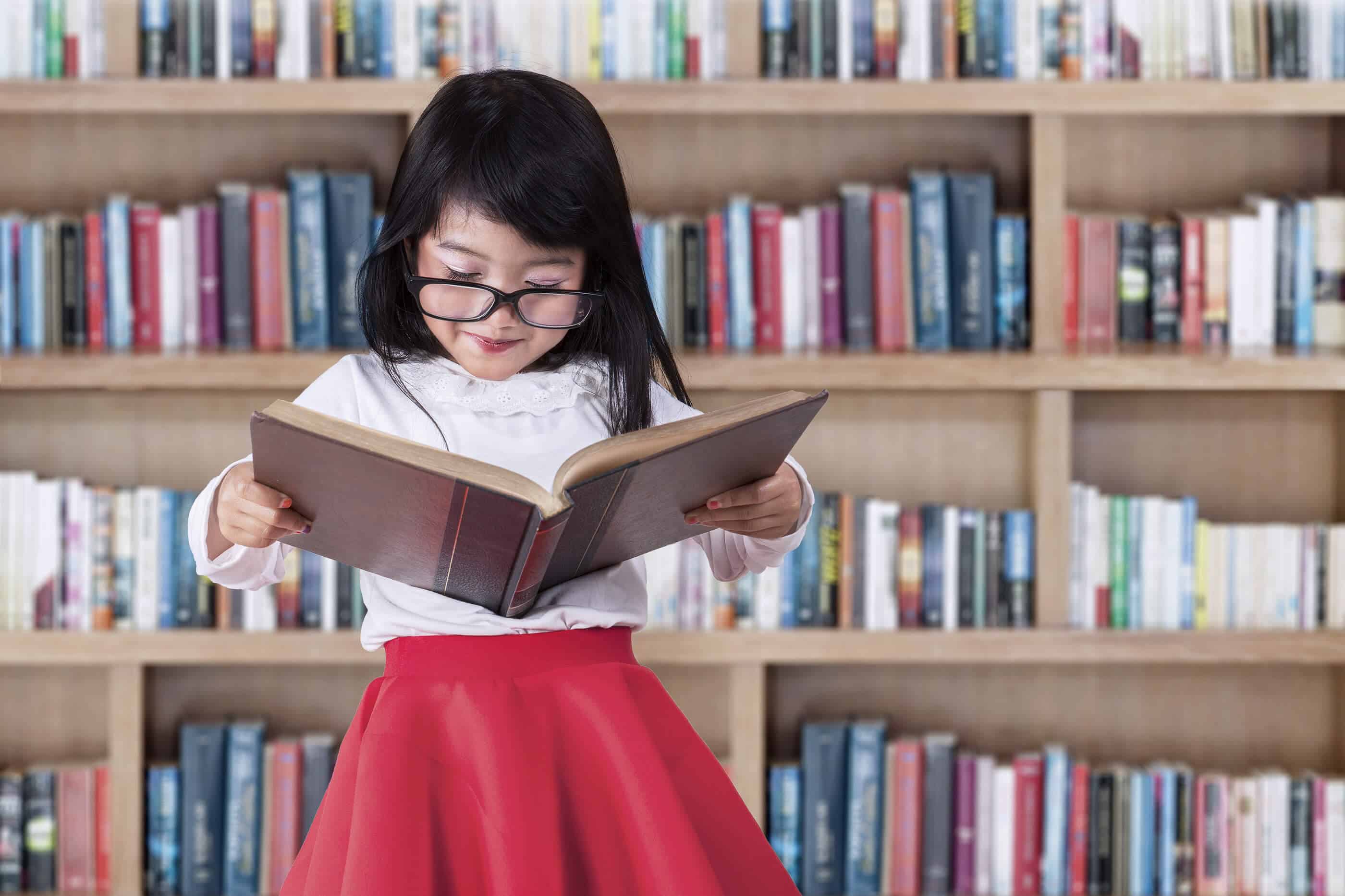 A young Asian girl is standing while holding a book that she is reading. A bookshelf full of books is behind her.
