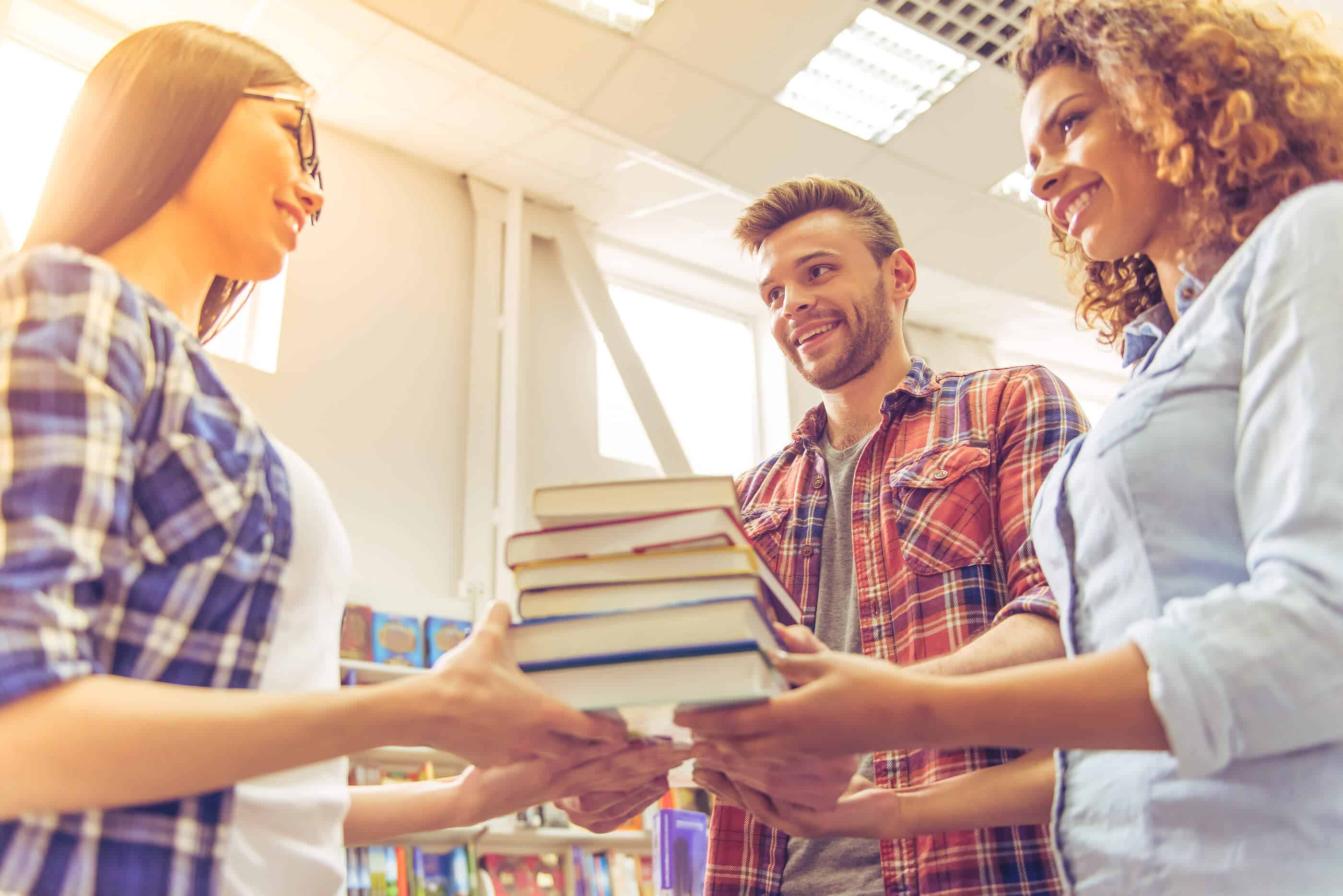 A group of three young people (two women, one man) is standing in a circle, all of them holding a stack of books in the center of them. The background is bright, but books on bookshelves can been seen.