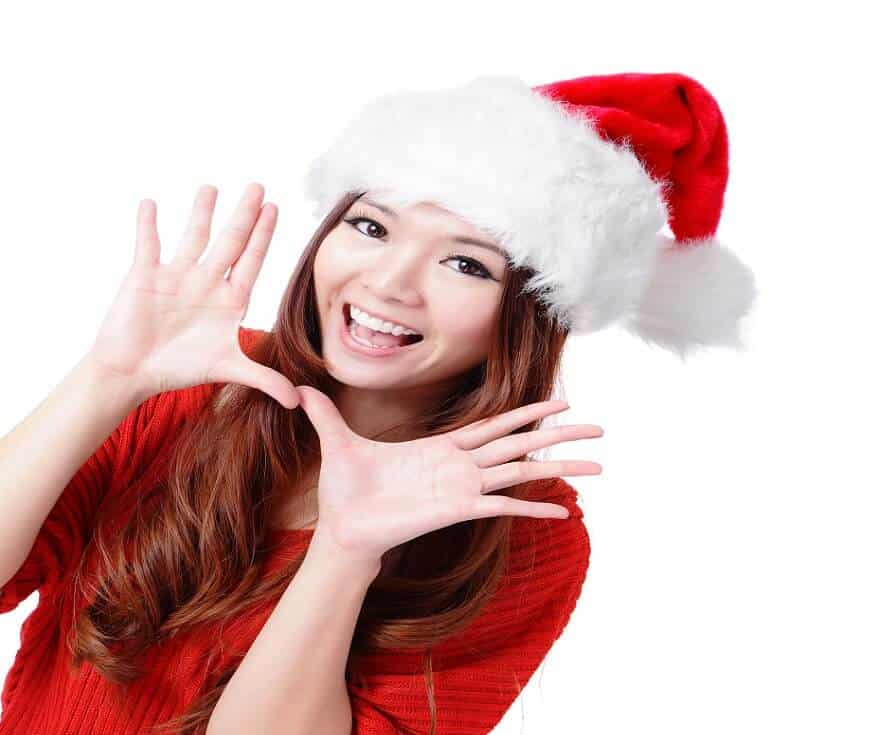 A young Asian girl dressed in a red santa outfit with both hands open waving "hello."