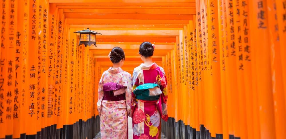 Torii Gates at Fushimi Inari Shrine. Two women dressed in traditional Japanese clothes can be seen walking through it side-by-side.