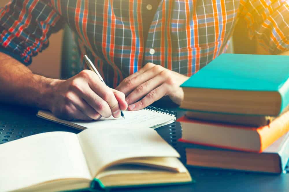 A close up of a man writing something in a notebook with a stack of books next to him.