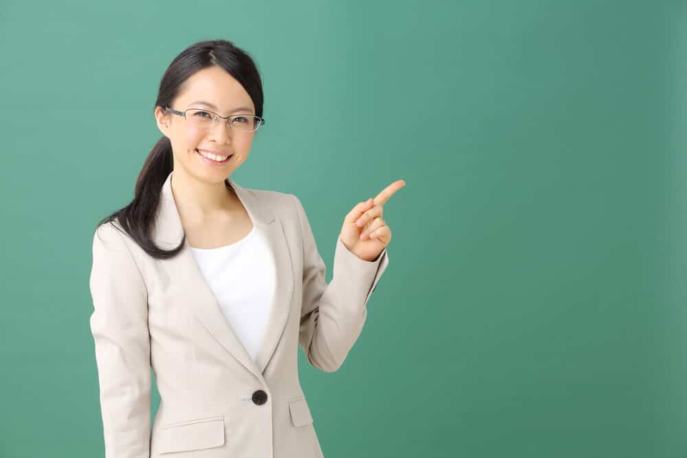 A young Asian woman wearing a greyish business suit and glasses smiling and pointing at something with her left pointer finger.