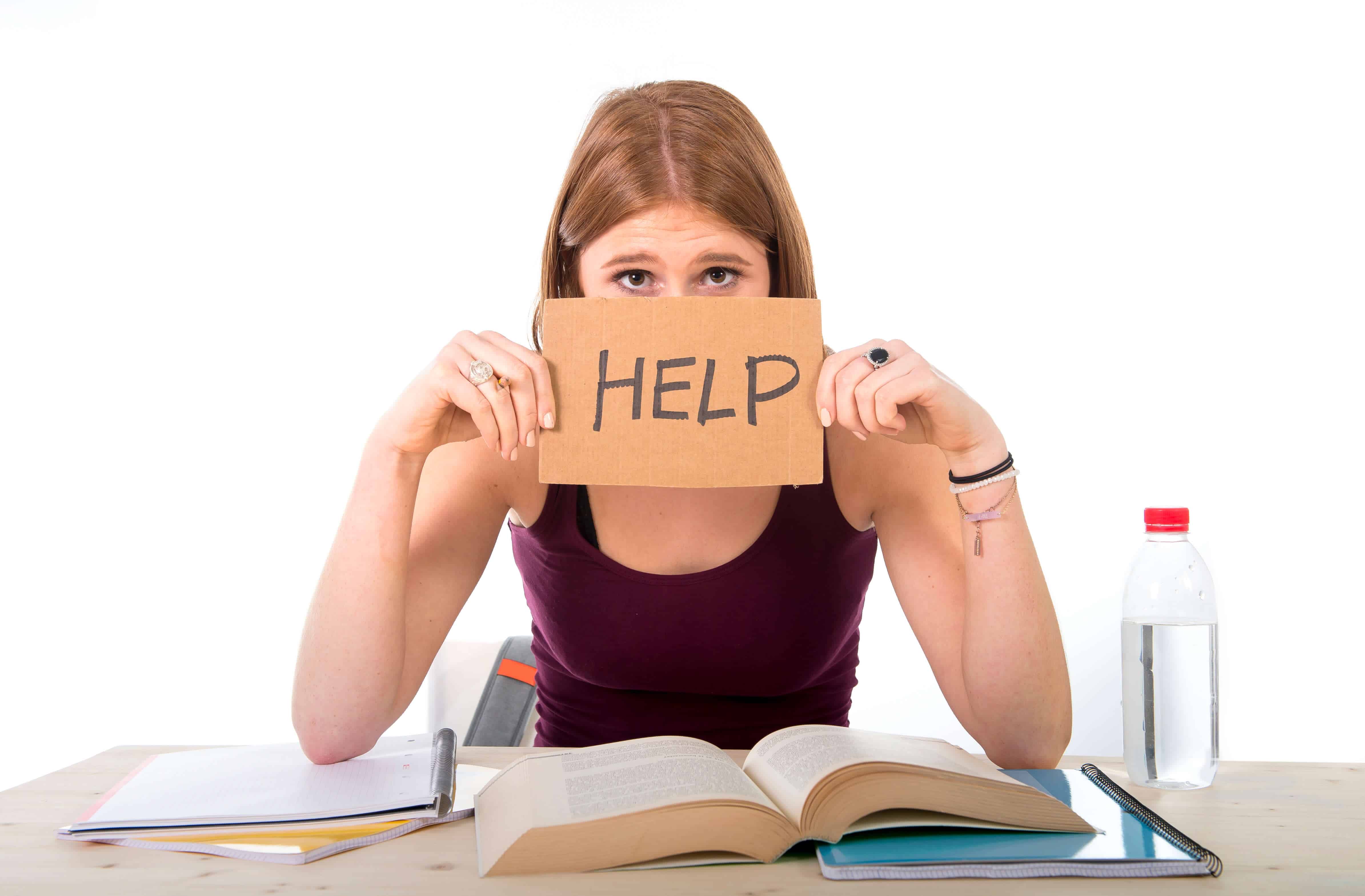 A young woman with dirty blonde hair is sitting down at a desk with an open book. She is hold a sign in front of her face that reads, "HELP."