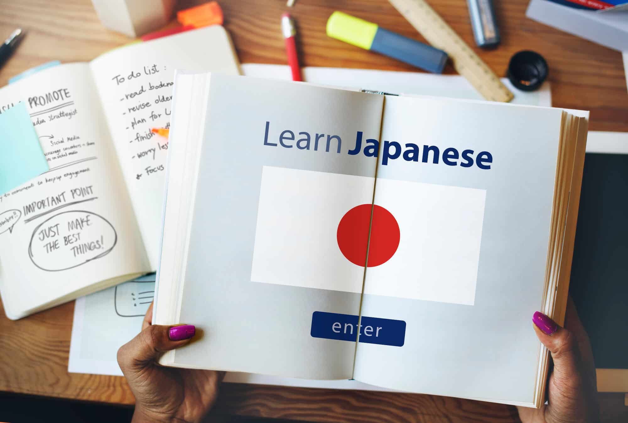 A woman's hands are holding a book open. Inside the book is a flag of Japan with a blue "enter" button and the text, "Learn Japanese" written at the top. In the background is a desk with other notebooks, pens, rulers, and a tablet on it.