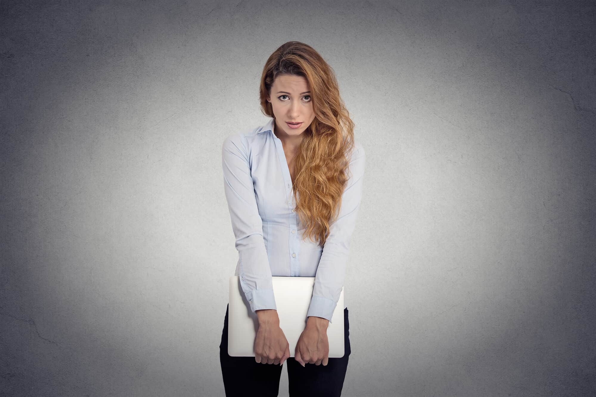 A young woman with long brown hair is carrying a laptop/note in her hands in front of her. She looks scared or shy.