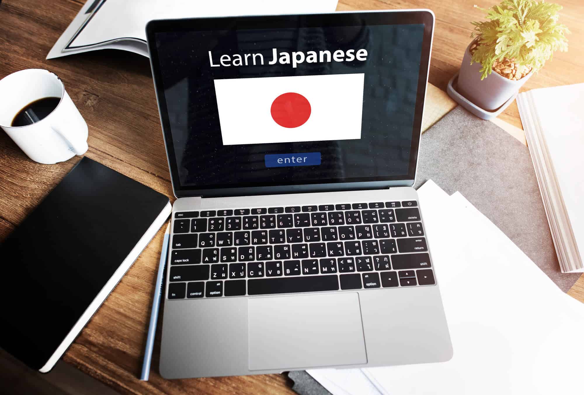 A laptop on a desk that is displaying a flag of Japan with a blue enter button at the bottom and a the text, "Learn Japanese" at the top. A coffee cup, notebook, notes, and a plant can also be seen on the desk.