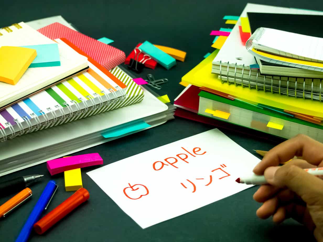 Two stacks of notebooks and folders with a bunch of color mini Post-It notes scattered all around. The the center is a white flashcard with the word, "apple" written on it in red. There is also a hand-drawn picture of an apple and Japanese characters on the same flashcard, also in red. Someone's hand can be seen holding a red pen.