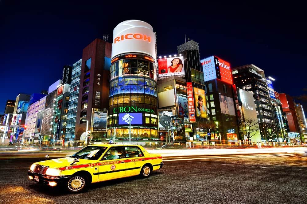 A taxi driving on the road in the foreground, with a lot of buildings and shops in the background in Ginza, Japan.