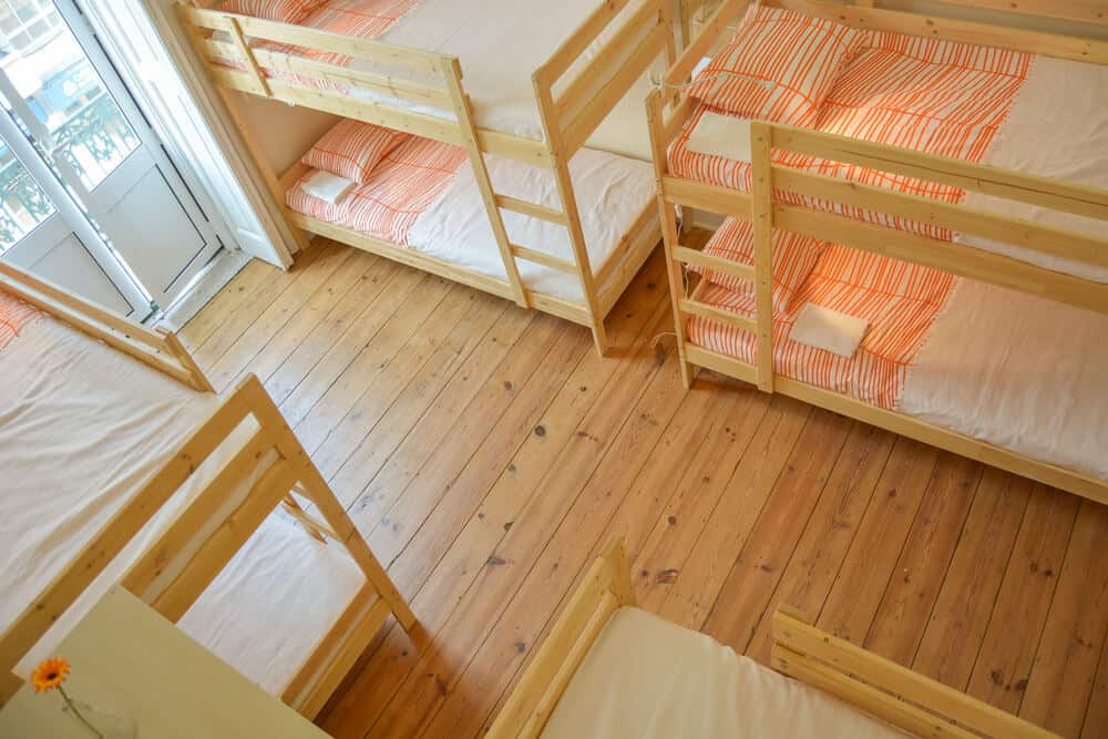 Four bunk beds with white and orange stripped sheets and pillows inside of a room in a hostel. The room is bright with sunlight coming through the door.