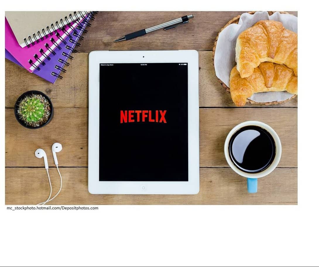 A smart tablet on a wooden table displaying th Netflix icon. Around the tablet are spiral notebooks, a smal cactus plant, earbuds, a pen, a basket with croissants in it, and a cup of coffee.