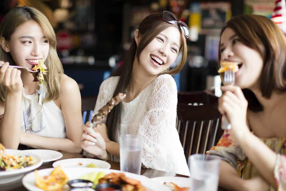 Three young, Asian women are sitting down at a table eating food and laughing.