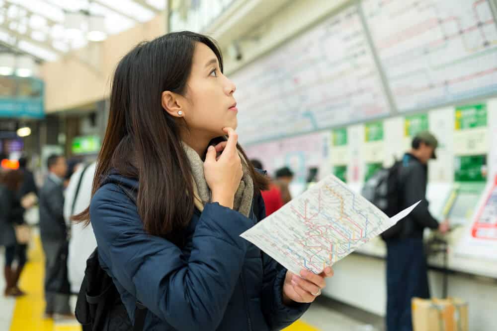 A young, Asian woman looks confused as she is holding ma and looking at a train ticket guide in Japan.