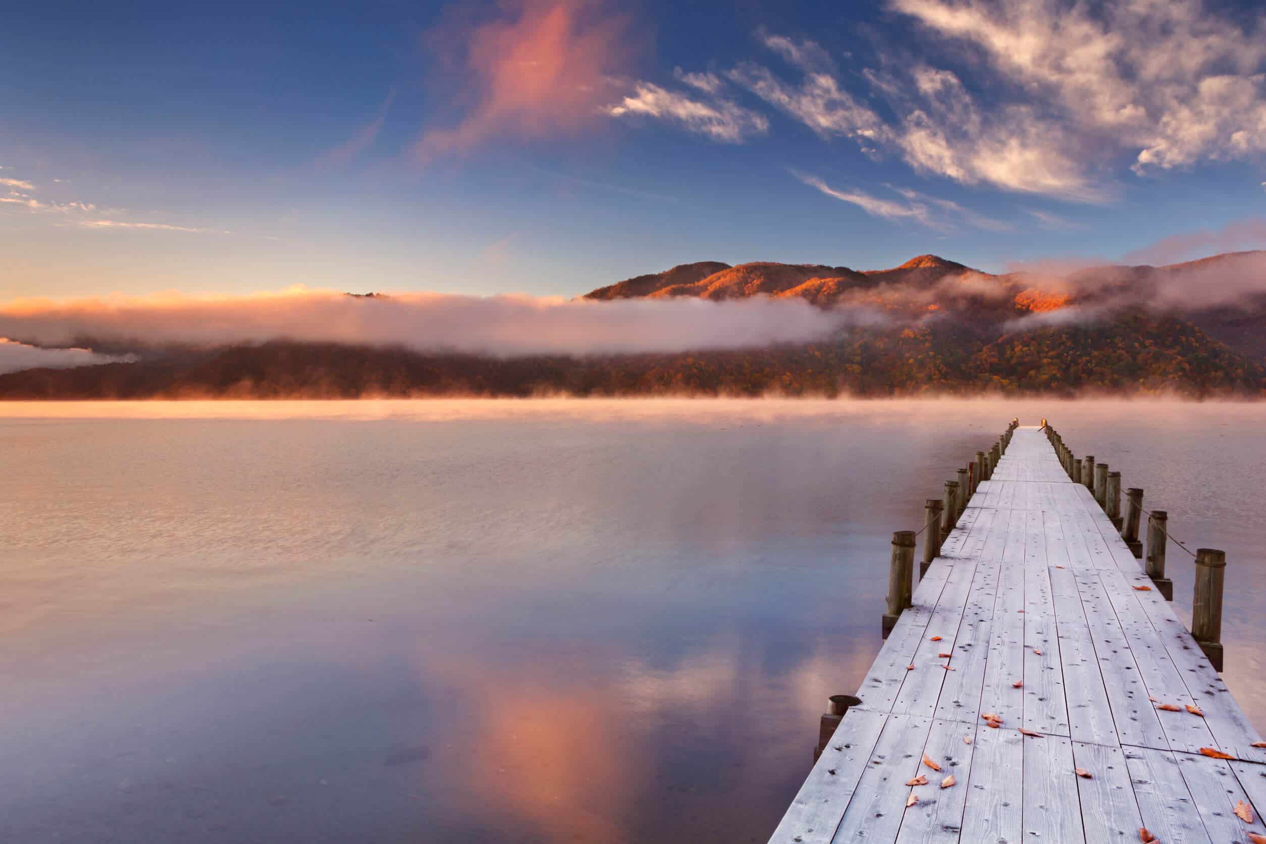 A wooden dock going over a calm lake. In the background, mountains with clouds over them can be seen.