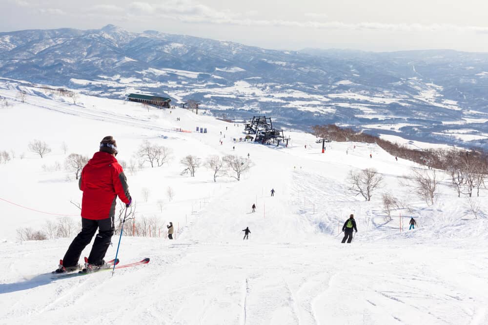 Skiiers are going down a hill that has a few trees without any leaves on it. In the background, a ski lift and a building can be seen, as well as snow on the ground and mountains in the distance.