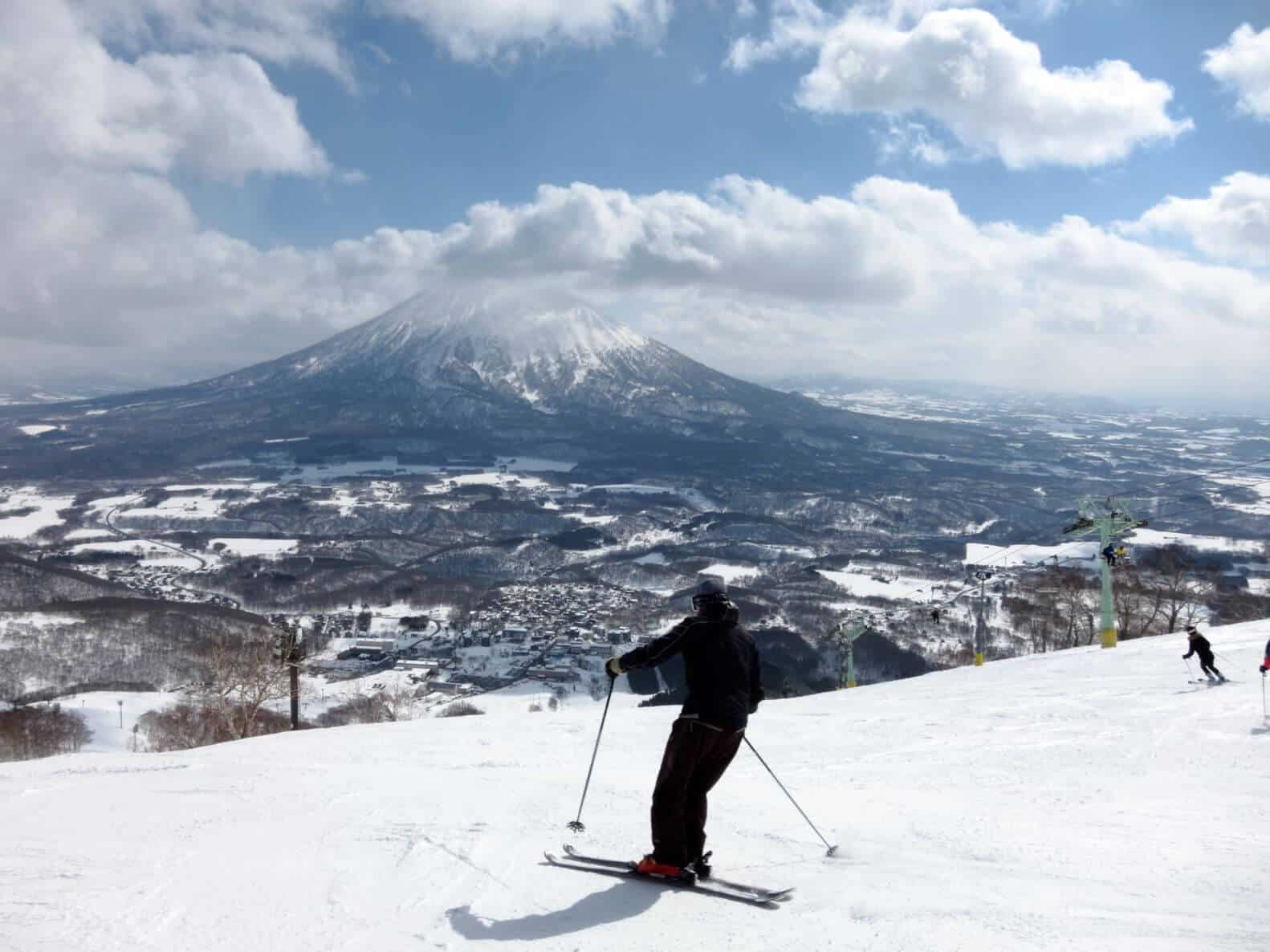 People skiing atop a high hill overlooking buildings and the ground below. A snow covered mountain is in the background.