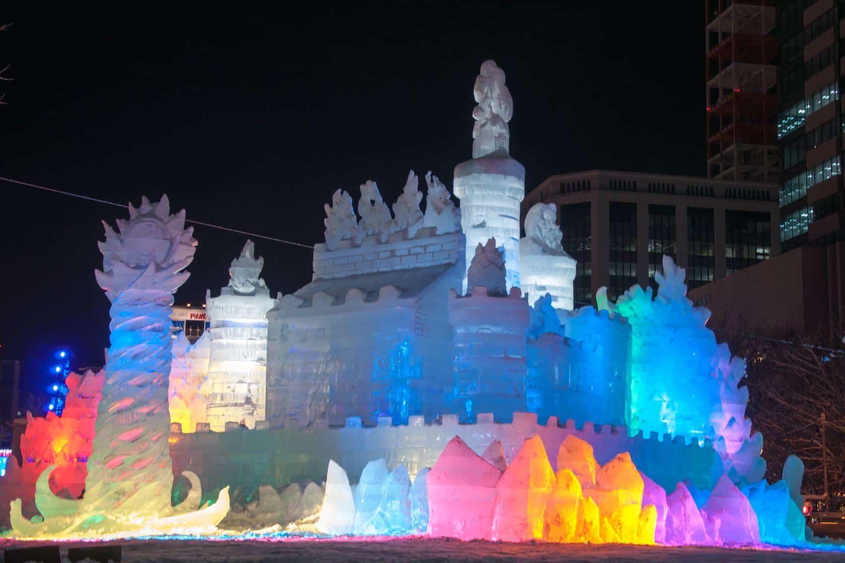 A huge snow sculpture of a big building with pillars around. At the bottom of this sculpture are ice pieces that have been illuminated with different colors (blue, pink, yellow).
