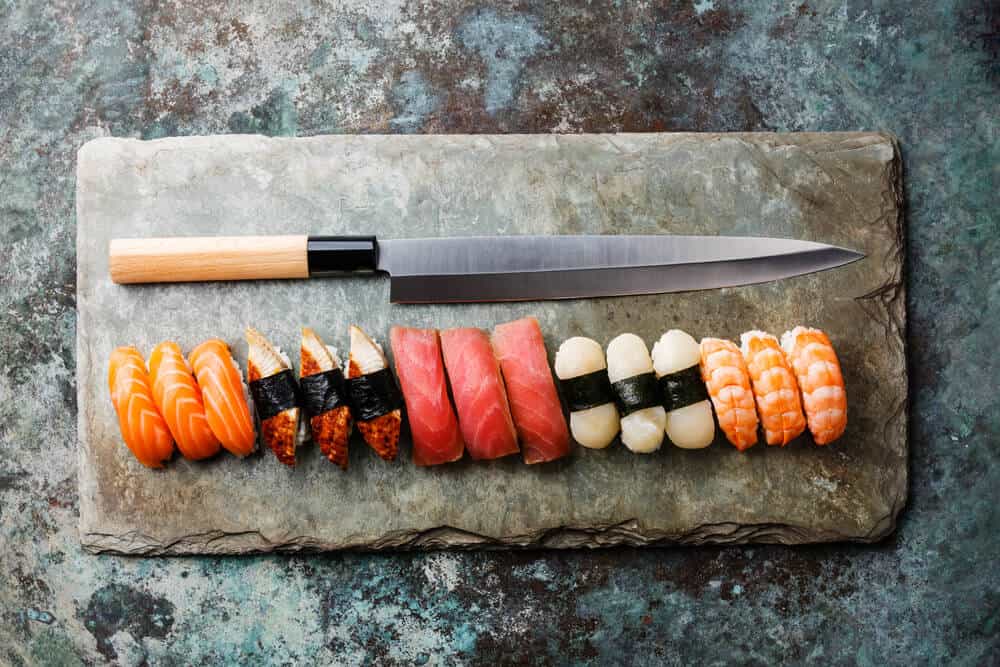 A long kitchen knife on a stone slab. In front of it is a row of nigiri sushi (salmon, unagi, maguro, hotate, ebi).