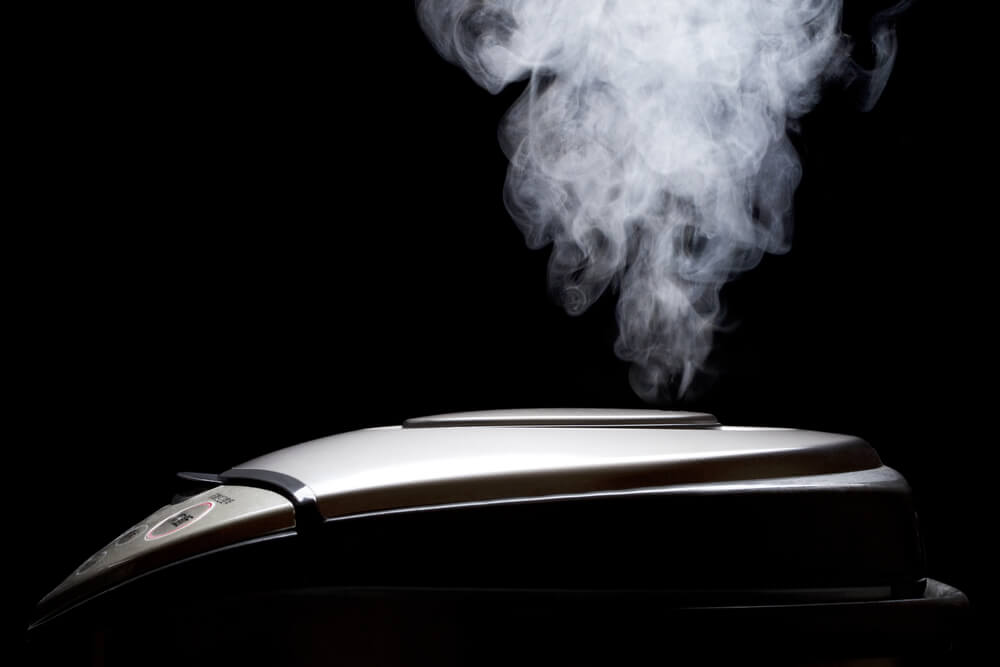A black and silver-ish rice cooker with steam coming out from the top.