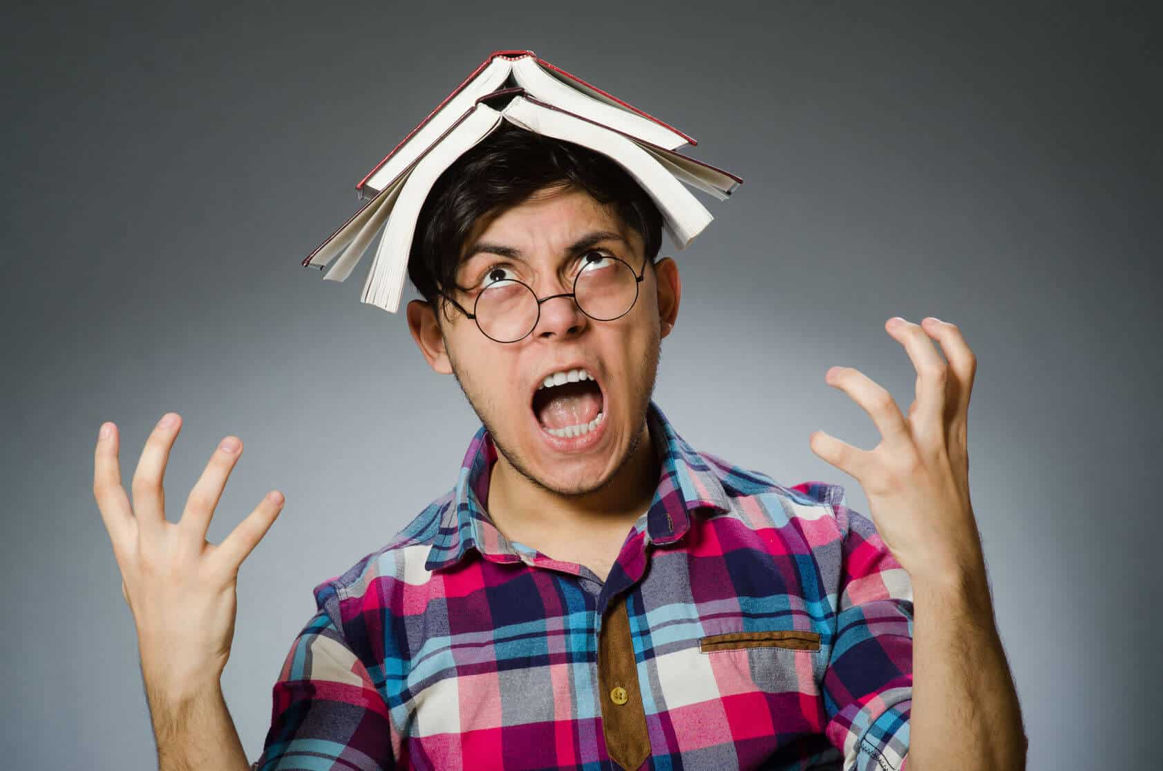 A young man with glasses has two open books on top of his head. He is looking up at them and screamign with both of his hands up.