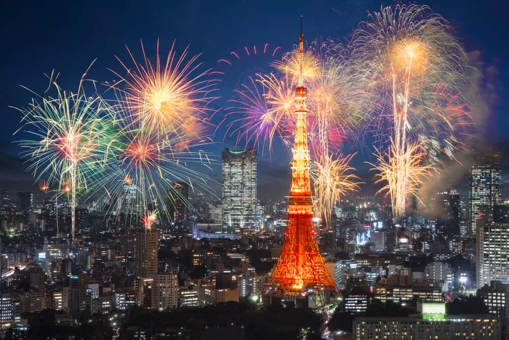 An image of fireworks in the sky with the many buildings of Tokyo all around. Tokyo Tower is in the center of this image.