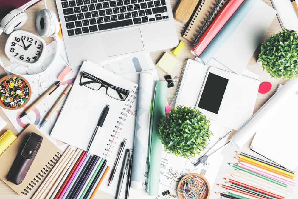 A desk that has notebooks, colored pencies, pens, a laptop and smarltphone, headphones, a clock, push pins, a plant, paper clips, and other office supplies spread out on top of a desk.