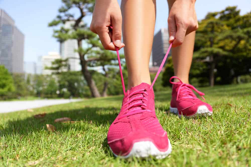 A woman is seen tying the laces of her pink shoes, outdoors. Trees and buildings can be seen blurred in the background.