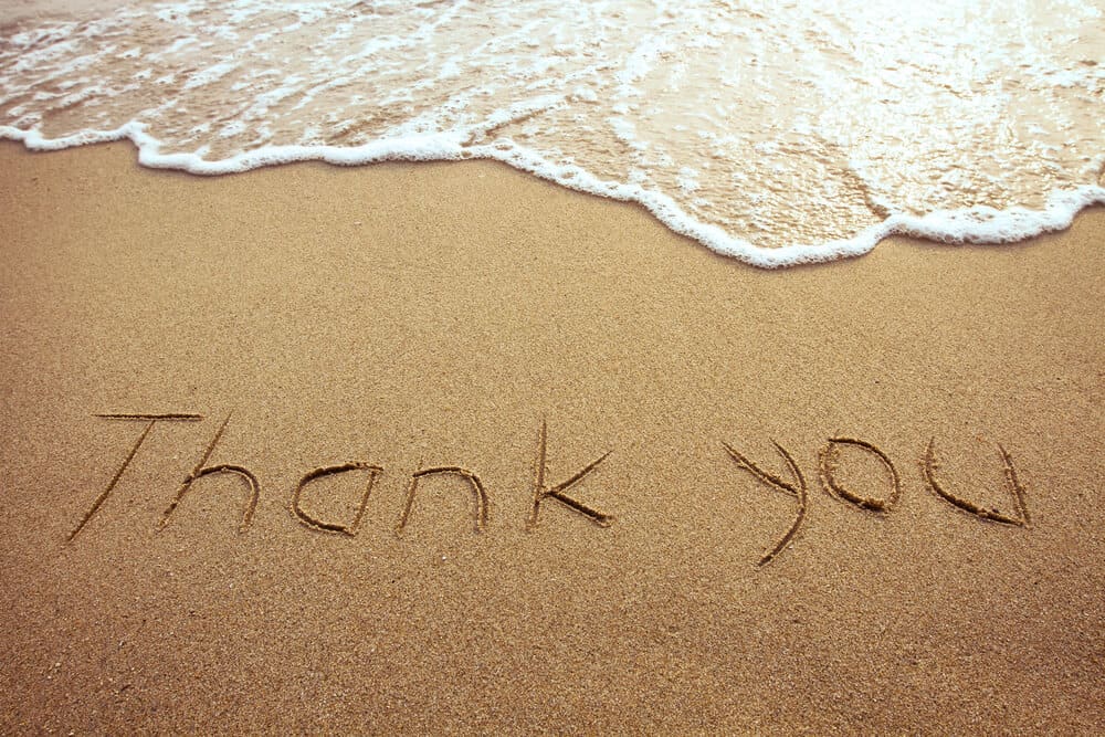 The words "Thank you" are written in the sand on a beach, with a small wave approachingit.