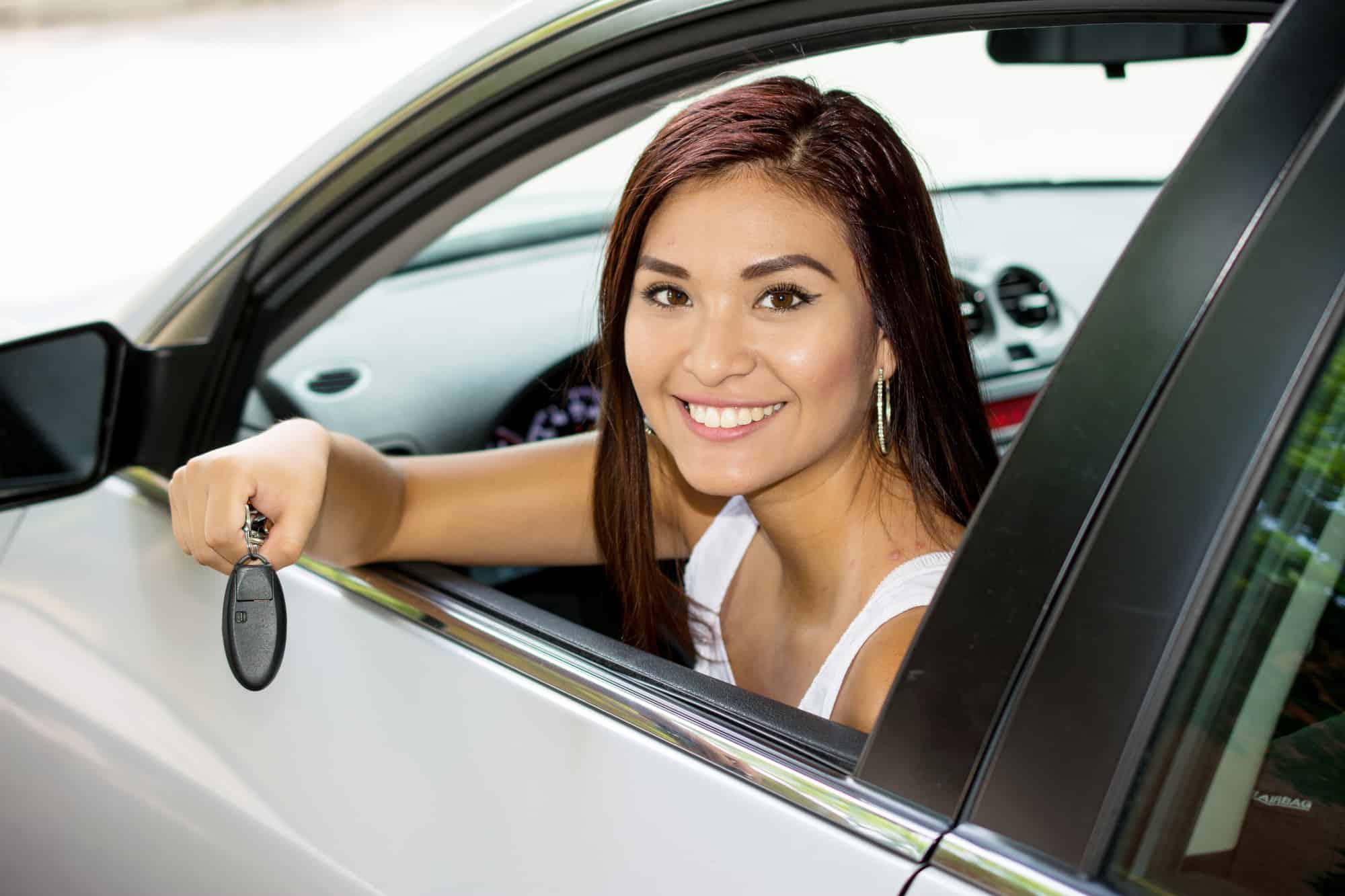 A young woman sitting in a car and looking out of the window and smiling. She is holding keys in her right hand.