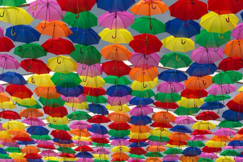 Dozens of umbrellas strung together above (maybe on the ceiling of a building) in all different colors like red, blue, yellow, green, orange, and pink. The picture is taken below the umbrellas.