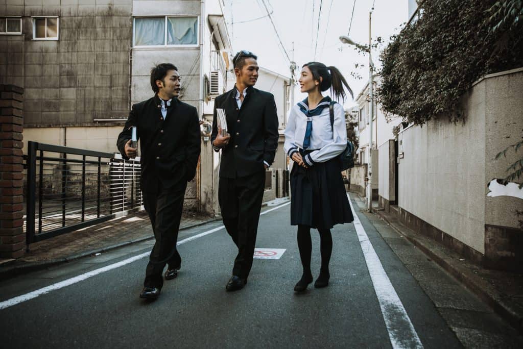 Two young boys and a young girl dressed in a Japanese school uniform. They are talking while walking down a residential road
