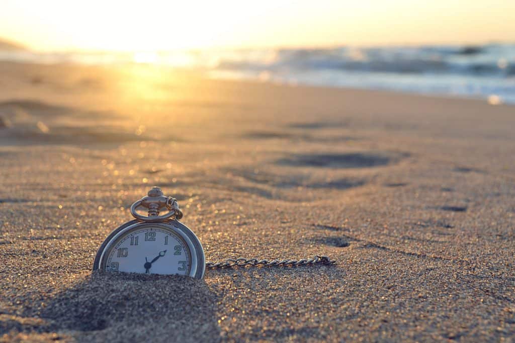 An older, metal stopwatch halfway embedded in the sand on a beach with the sun setting in the backgroun