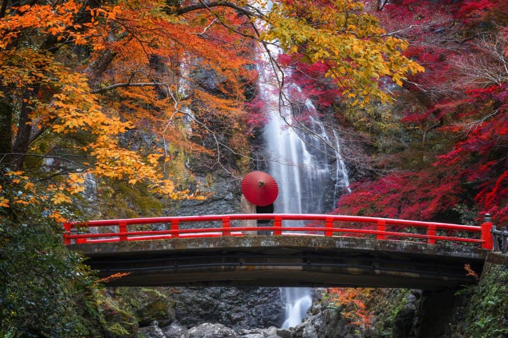 Minoo Park Waterfall with a couple under a red Japanese style umbrella on a red bridge in front of the waterfall.