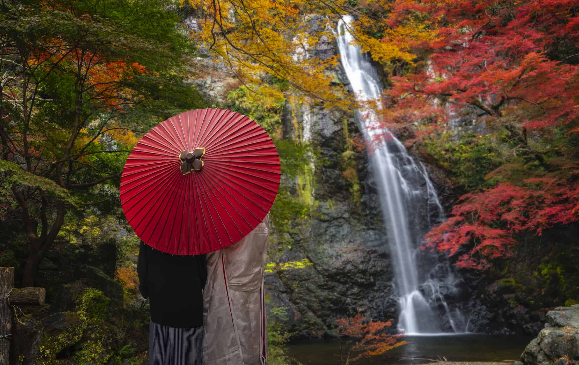 Minoo Park Waterfall in Osaka, Japan. A couple is standing in front of the waterfall under a red, Japanese style umbrella.