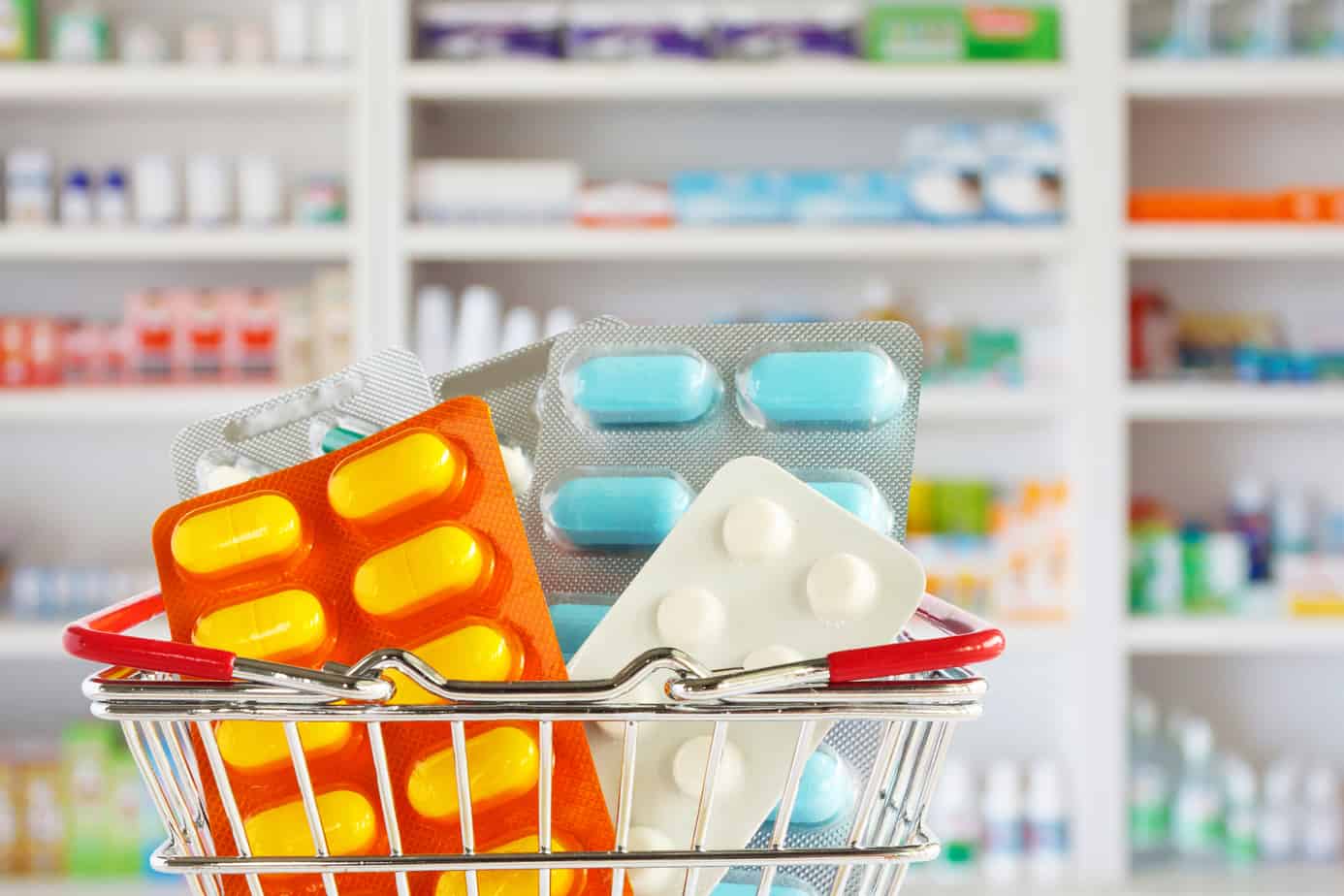 Medicine pill tablets in their pill packs, which is in a mini shopping cart with a blurred view of a pharmacy drugstore's shelves in the background.