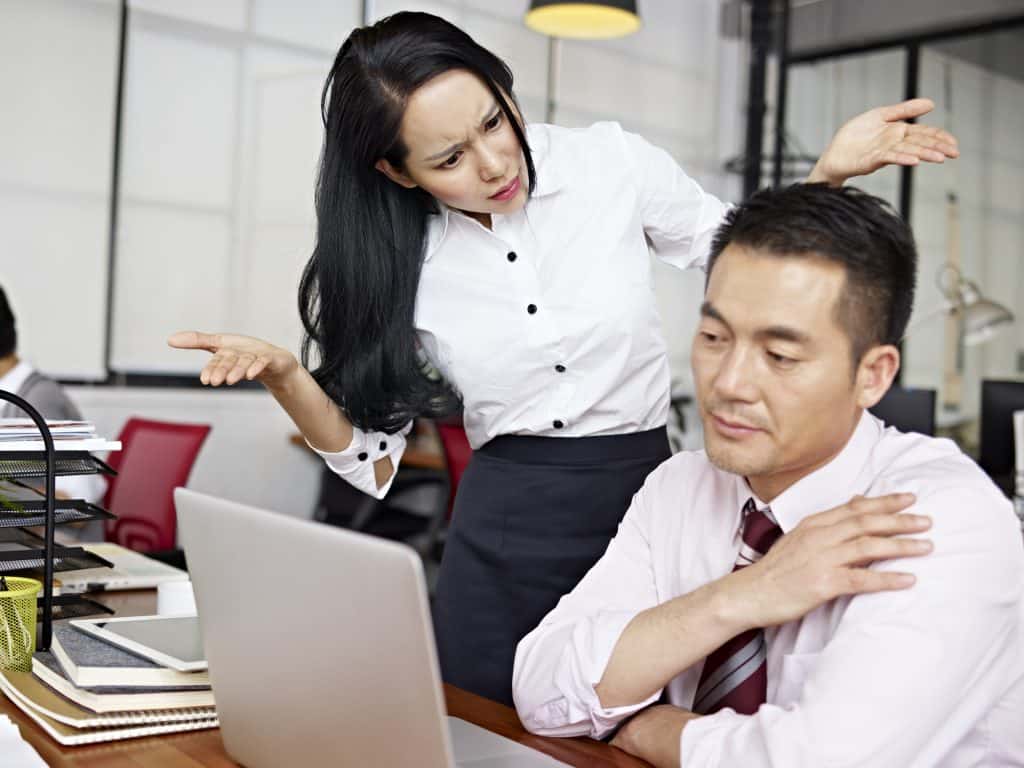 Asian male sitting at his desk working on his laptop while a young asian woman is shrugging her shoulders looking angrily at him