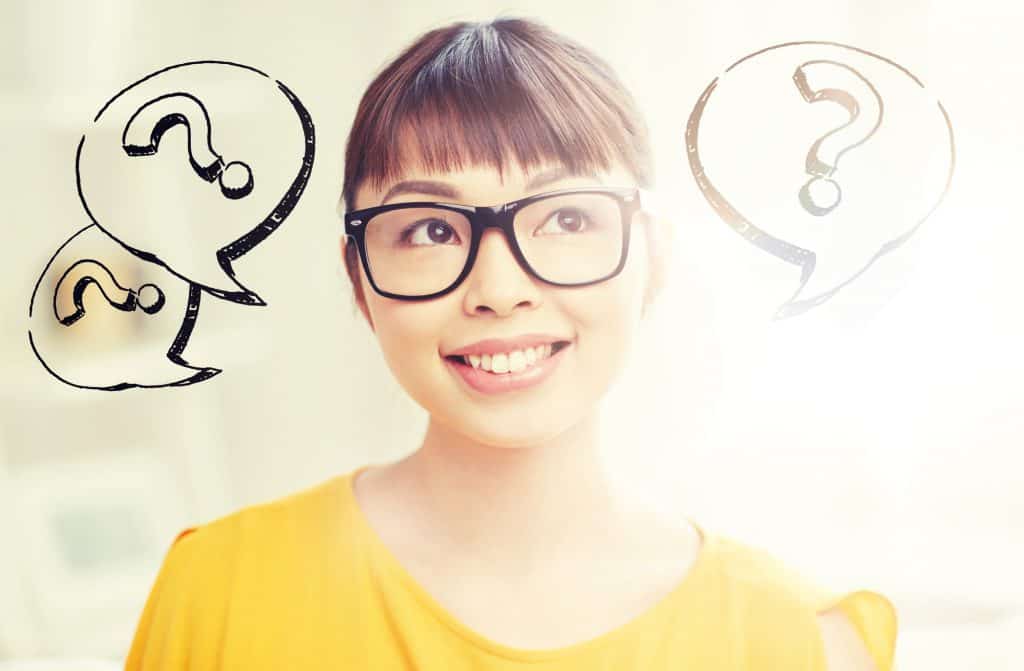 Young Asian woman with glasses wearing a yellow top smiling but with a slight confused look on her face. Question marks in speech bubbles are surrounding her head