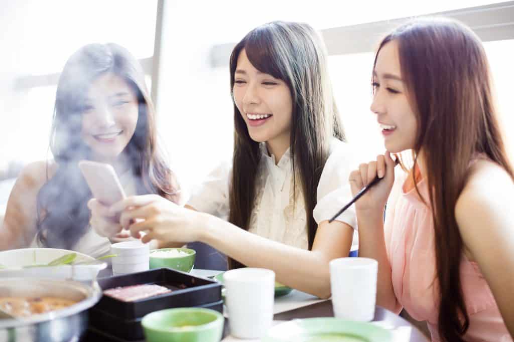 Three young Asian women sitting at a table eating hot pot. The woman in the middle is holding a smartphone, which all three of them are looking at and smiling.