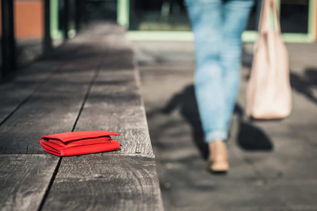 A woman wearing jeans hold a beige bang is seen in the background, walking away from a red wallet left on a bench.