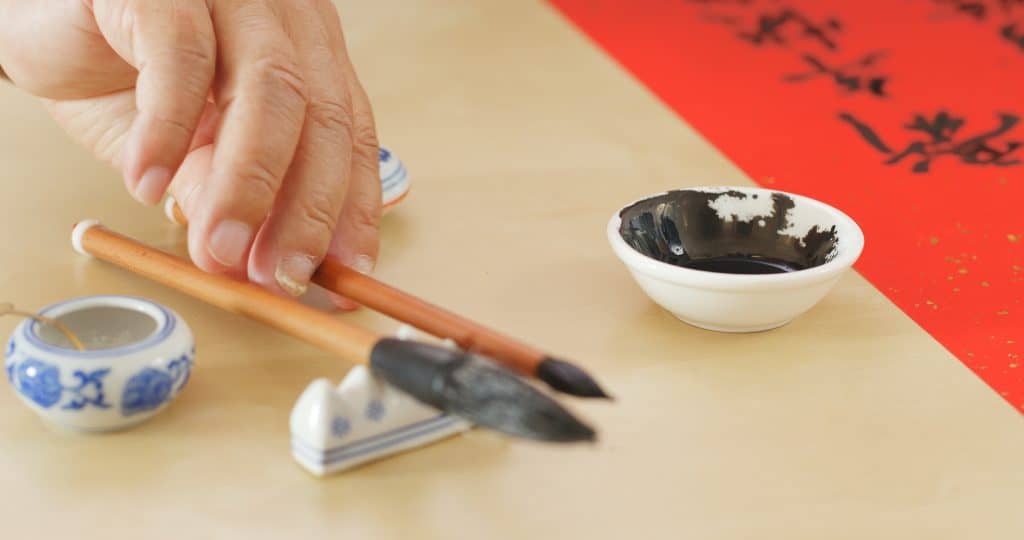 A picture of someone's hand about to take one of two calligraphy brushes resting on a small stand on the table. In the backgroud there is a small white dish filled with black ink and a red paper with Chinese characters written on it.