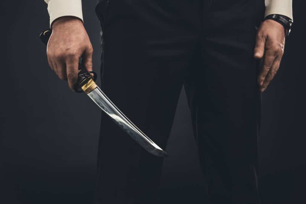 A man shot from the waist down. He is wearing a white long sleeve shirt and black pants, and he is holding an samurai style of short sword in his right hand.