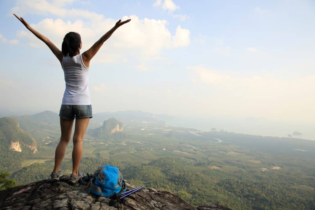 A girl on the edge of a cliff overlooking the green nature and a body of water below. The girl is wearing a white tank top and jean shorts, and has both of her hands raise up above her head.