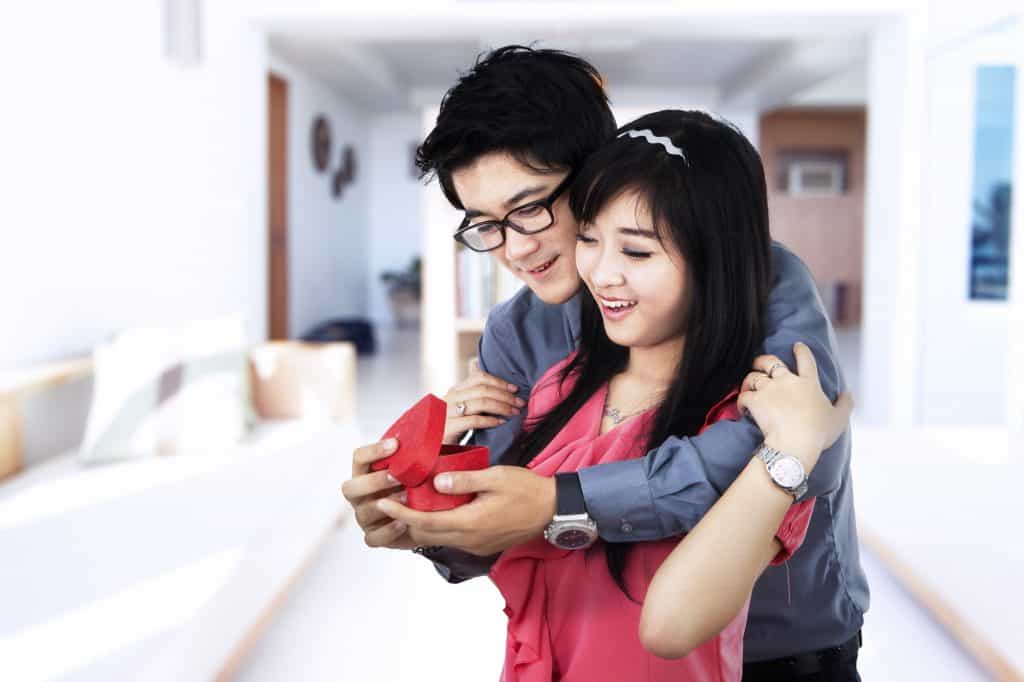 A young Asian couple. The man is embracing the woman from behind, holding and showing her a ring in a red box. The woman is holding the man on his shoulder, while smiling at the ring. The woman is wearing a pink outfit and the man is wearing a long sleeve grey shirt.