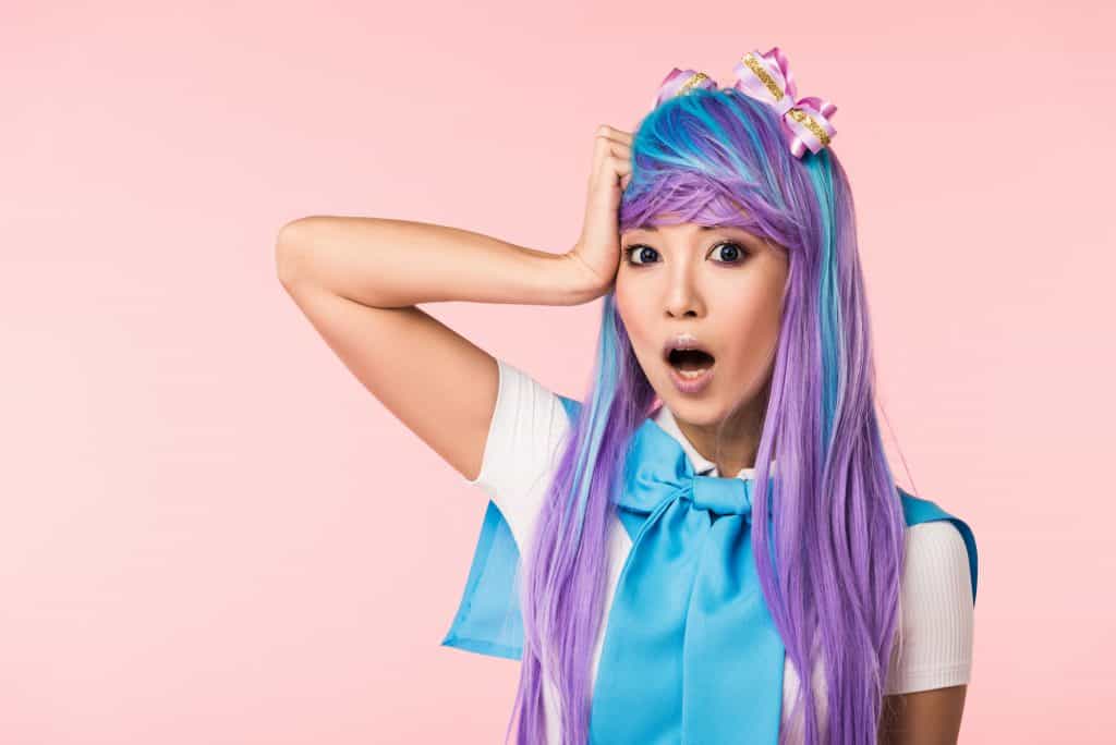 A young Asian girl with long purple and blue hair, dressed in a white and blue outfit. The blue part of her outfit has a bow tie by her neck. She has her right hand on the side of her right temple, with her mouth open to express shock or surprise.