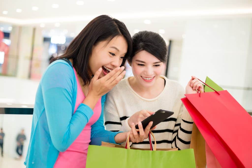 Two young Asian woman looking at a cellphone while holding a bunch of green and pink shopping bags. They looked suprised.